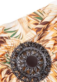 Blumengemusterter Stoff in Orange, Creme und Grün, mit einer metallischen silbernen Brosche mit filigranen Details in der Mitte.