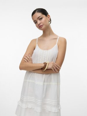 Witte, mouwloze jurk met delicate verticale borduursels en een gerimpelde zoom. Model draagt houten en doorzichtige armbanden. Eenvoudige achtergrond.