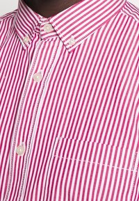 Chemise boutonnée avec des rayures verticales rouges et blanches, dotée d'un col, d'une poche avant et de boutons clairs. Texture en coton lisse.