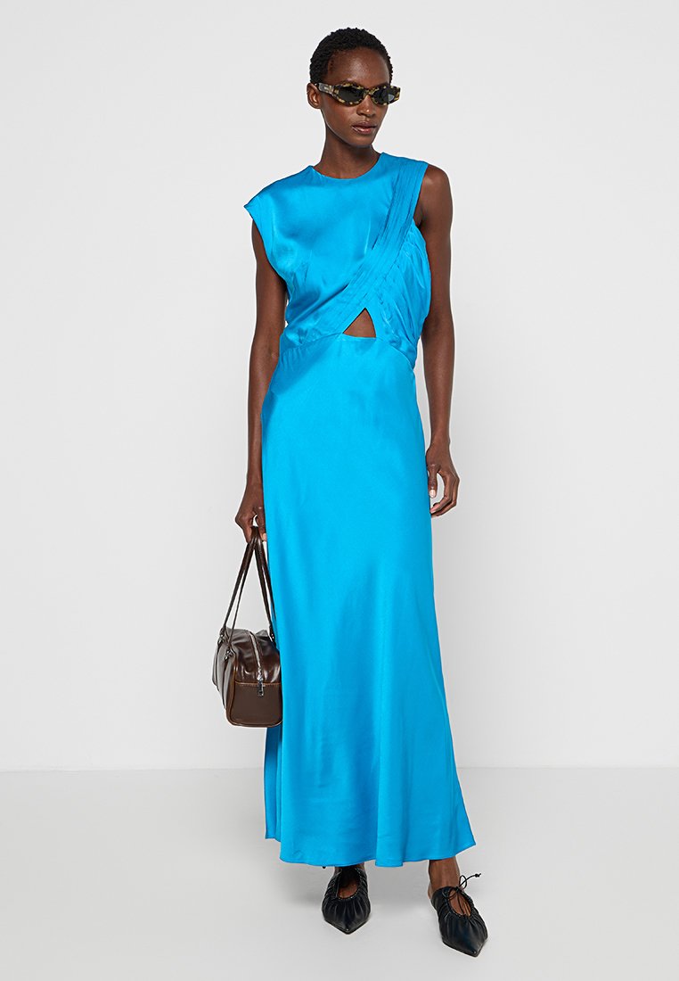 Abito maxi in satin blu con scollatura senza maniche, caratterizzato da un design torciglione e una apertura laterale. Texture morbida, abbinato a una borsa marrone e scarpe nere.