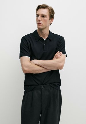 Jeune homme aux cheveux châtain clair portant un polo noir à manches courtes et un pantalon foncé, les bras croisés, regardant sur le côté sur un fond blanc uni.