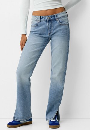 Kobieta w jasnoniebieskich prostych jeansach z wysokim stanem, bocznymi rozcięciami i niebieskich trampkach, zestawiona z białym długim rękawem crop top.