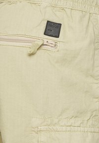Vaaleankeltaiset cargo-shortsit, jotka on valmistettu teksturoidusta kankaasta. Niissä on vetoketjullinen tasku ja musta "Alpha Industries" -logopatchi.