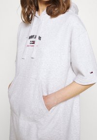 Šedá oversized mikina s kapucí, šňůrkami, přední kapsou a detaily loga. Vyrobena z měkké tkaniny, má krátké rukávy a uvolněný střih.