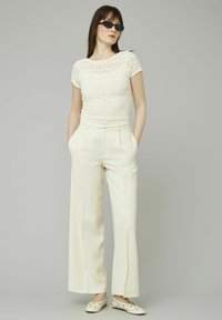 Haut blanc texturé en dentelle avec des manches courtes, pantalon large taille haute crème, et chaussures blanches décoratives à plat avec des clous.