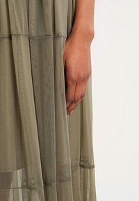 Vestido de capas translúcidas en verde oliva con textura fluida. Presenta un ajuste holgado y una cintura fruncida, mostrando una mano sosteniendo la tela.