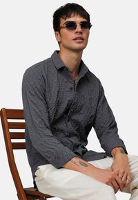 Camisa de manga larga gris con textura, cuello abotonado y detalles de costura visibles, combinada con pantalones de color claro, sentada en una silla de madera.