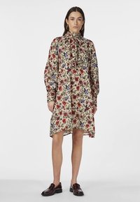 Cvjetna midi haljina s dugim rukavima, kremasta osnova, uzorak crvenih i plavih cvjetova, detalj sa vezanjem na vratu i asimetričan rub. Nošena s tamnim mokasinama.