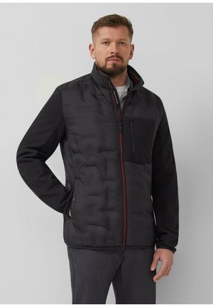 Mann steht mit Händen in den Taschen, trägt eine schwarze gesteppte Jacke mit Reißverschlusstasche auf der Brust und graue Hose vor grauem Hintergrund.