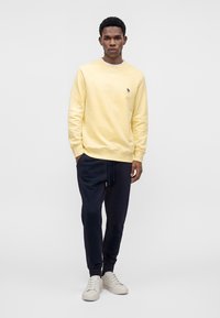 Homme debout portant un sweat-shirt jaune clair, un pantalon de jogging bleu marine foncé et des baskets blanches sur un fond clair uni.