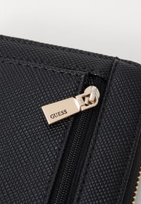 Must mustrikkidega must kunstnahast käekott, millel on kullast tõmblukk ja "GUESS" brändingu logo. Kotil on struktureeritud disain ja kontrastsed õmblused.