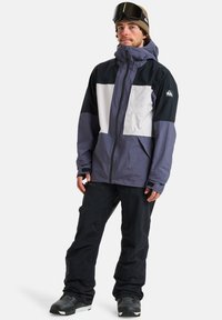 Skijacke mit wasserdichtem Design, ausgestattet mit schwarzen, weißen und lila Paneelen, verstellbarer Kapuze und Reißverschluss vorne. Ergänzt durch schwarze Hose.