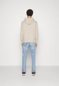 Sudadera de color beige claro con capucha, puños y dobladillo acanalados, combinada con jeans sueltos de color azul claro y zapatillas oscuras. Los detalles incluyen una textura suave.