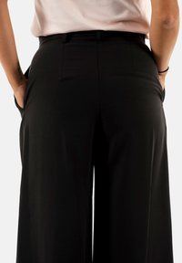 Pantalones de pierna ancha negros con textura suave, que cuentan con bolsillos laterales y una cintura ajustada. La vista trasera muestra costuras confeccionadas y un acabado limpio.