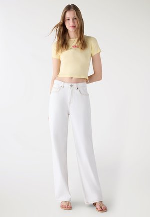 Giovane donna in piedi che indossa una t-shirt gialla corta con dettaglio floreale, jeans bianchi a vita alta e gamba larga, e sandali su sfondo bianco.