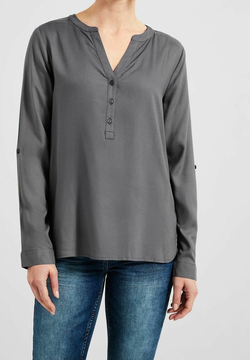 Blouse grise à manches longues avec trois boutons devant, portée avec un jean bleu, montrée sur une personne du cou aux hanches sur un fond uni.
