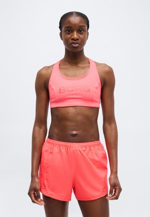 Femme debout, portant un brassard de sport corail et un short de sport assorti, face à l'avant sur un fond clair uni.