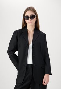 Svart blazer med én lomme, ensfarget design og to knapper. Brukt over en hvit topp, kombinert med svarte bukser og solbriller.