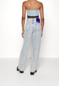 Mujer que lleva unos pantalones vaqueros rectos de cintura alta en azul claro con una etiqueta azul en el bolsillo trasero y un top corto de mezclilla sin mangas.