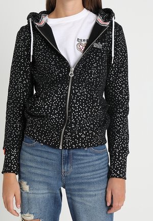 Femme portant un sweat à capuche noir zippé avec des motifs d'étoiles blanches, un T-shirt blanc avec un motif pizza, et un jean bleu déchiré.