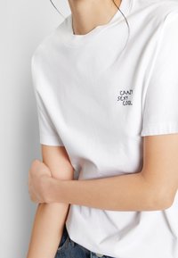 Osoba v bílém tričku s vyšitým nápisem "CRAZY SEXY COOL" na hrudi, která drží levou ruku pravou.