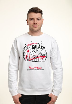 Mężczyzna w białej bluzie z nadrukiem logo „Pizza Planet” w kolorach czerwonym i czarnym oraz napisem „Najświeższe w Galaktyce”, stojący z rękami w kieszeniach.