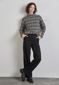 Femme aux cheveux bouclés portant un haut noir et blanc à manches longues à motif, un pantalon large noir et des chaussures noires, debout contre un mur gris.