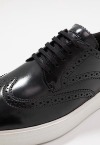 Chaussure en cuir noir avec des détails perforés, une finition lisse et des fermetures à lacets. La semelle est blanche avec un design minimaliste.