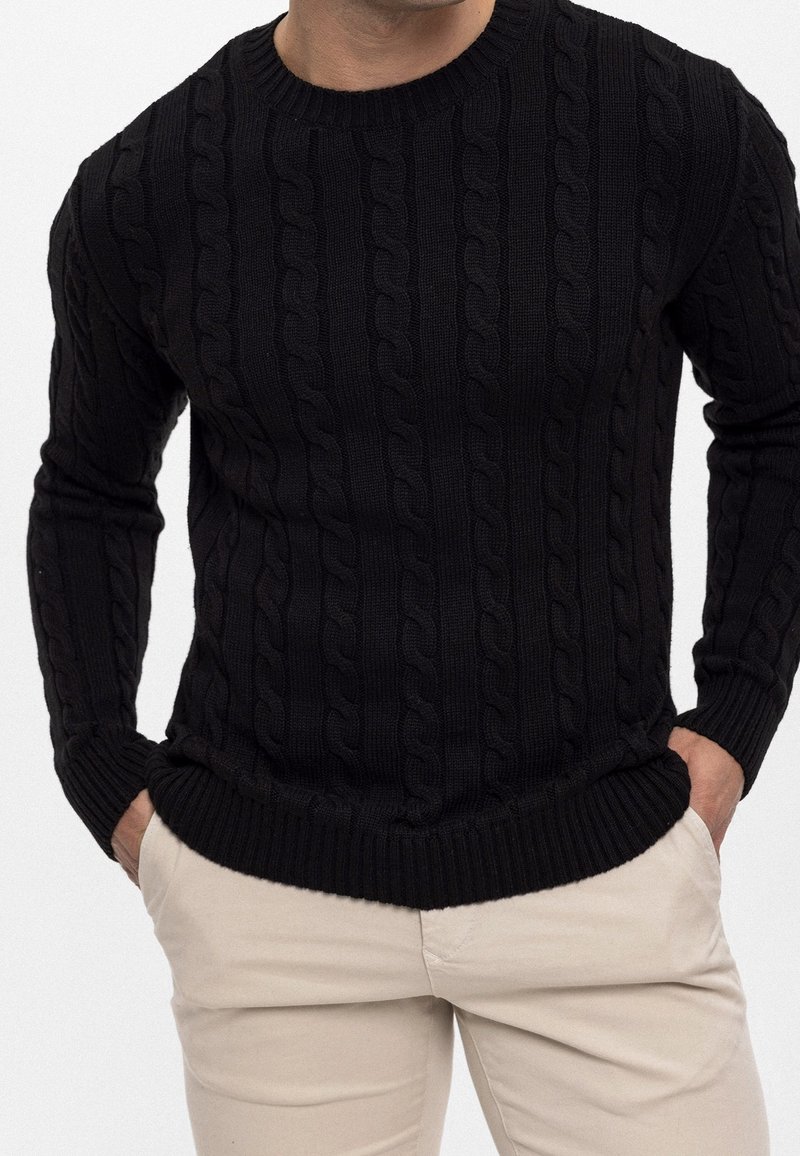 Pull noir en maille avec un motif torsadé en relief, col rond et poignets côtelés, coupe ajustée. Associé à un pantalon de couleur claire.