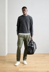 Sweatshirt gris avec col blanc, pantalon vert olive et baskets blanches. Sac de sport en cuir noir avec un détail de quincaillerie ronde. Fond en parquet.