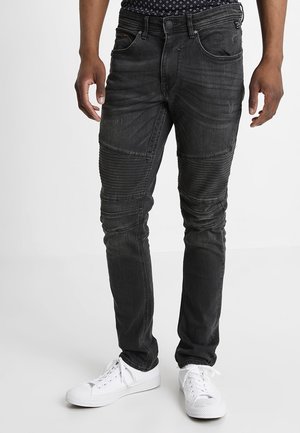 Uomo che indossa jeans slim-fit neri a coste con dettagli sulle ginocchia e sneakers bianche, in piedi contro uno sfondo chiaro e uniforme.