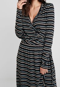 Zwarte wikkeljurk met lange mouwen, voorzien van horizontale strepen in blauw, crème en zwart. De stof lijkt zacht en rekbaar, met een elastische taille.