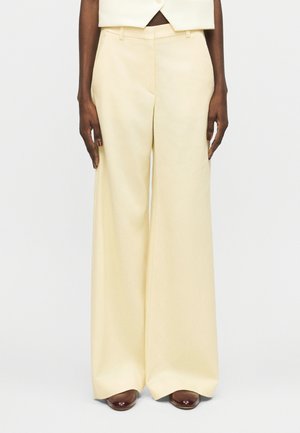 Εκρού παντελόνια φαρδιάς γραμμής φορεμένα με καφέ παπούτσια, κοντό εκρού σακάκι ορατό στη μέση, χέρια χαλαρά δίπλα στο σώμα σε λευκό φόντο.