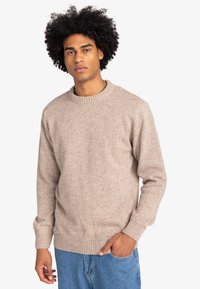 Jonge man met krullend haar, gekleed in een beige gebreid trui en blauwe spijkerbroek, staande met een hand in zijn pocket tegen een witte achtergrond.