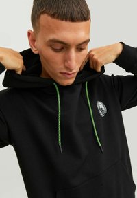 Sweatshirt noir avec une poche avant, cordons de serrage vert, et un logo circulaire sur la poitrine. Le tissu semble doux et texturé.