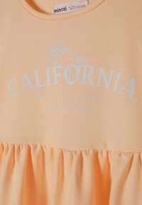 Różowa bawełniana sukienka z falbaną, z niebieskim nadrukiem tekstu "CALIFORNIA" oraz grafiką palm. Gładka tekstura, casualowy design.
