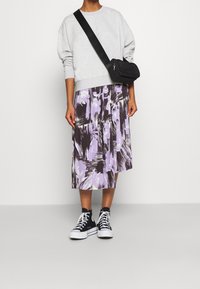 Felpa grigia, gonna midi a fantasia viola e nera, borsa a tracolla nera e sneakers alte nere. In piedi su uno sfondo neutro.