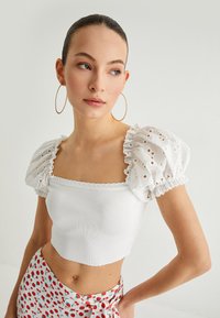 Weißes, kurzes Oberteil aus strukturiertem Rippmaterial mit puffärmel aus Spitze. Das Modell kombiniert es mit einem gemusterten Rock.