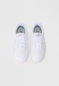 Pāris baltu Puma sporta apavu ar auklēm, zelta logo uz mēles un sāniem, skatīts no augšas uz balta fona.