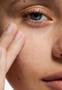 Gros plan sur une peau à la texture lisse, avec de légères taches de rousseur, un éclat naturel et un petit piercing au nez. Une main touche doucement la joue.