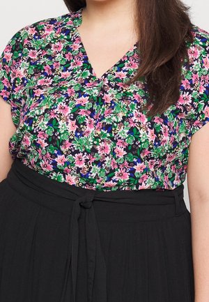 Kobieta w krótkim, kwiatowym bluzce z różowymi i zielonymi kwiatami oraz czarnej spódnicy z wysokim stanem, związanej kokardą w talii.