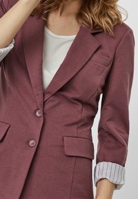 Wino-czerwony żakiet z dwoma guzikami, kieszenią na piersi oraz dwiema kieszeniami z przodu. Paskowe mankiety na podwiniętych rękawach wzbogacają design.