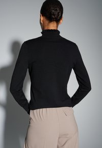 Haut noir à manches longues avec un col montant à volants et une silhouette ajustée, assorti à un pantalon beige clair avec un détail de poche arrière.