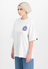 Biele premožené tričko vyrobené z bavlny, s fialovým grafickým nápisom "ALPHA INDUSTRIES" na prednej strane a malou nášivkou s logom na rukáve.