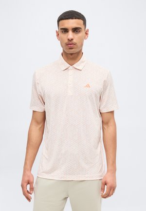 Muž oblečený v svetloružovej polo košeli s vzorom, golierom a zapínaním na gombíky, stojaci na jednoduchom bielom pozadí.