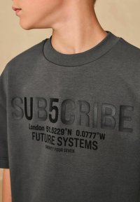 Tricou gri din bumbac, cu guler rotund texturat și inscripții rezemate de culoare neagră, inclusiv coordonate și "SISTEME DIN VIITOR" pe partea din față.