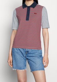 Polo rayé avec des rayures bordeaux et blanches, col bleu foncé, et boutons. Associé à un short en denim délavé clair avec ourlet effiloché.