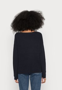 Pull en tricot marine avec un large col et des manches longues. Présente une texture côtelée et une coupe décontractée, associé à un jean en denim.