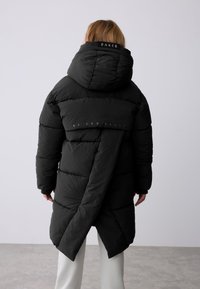 Zwarte pufferjas met een grote capuchon, voorzien van horizontale stiksels, een hoger-lager zoom en een logo op de achterkant. Gladde textuur en lichtgewicht.