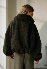 Dunkelgrüne, flauschige Fleecejacke mit hohem Kragen, gesenkten Schultern und elastischem Saum. Die strukturierte Oberfläche verleiht Tiefe.